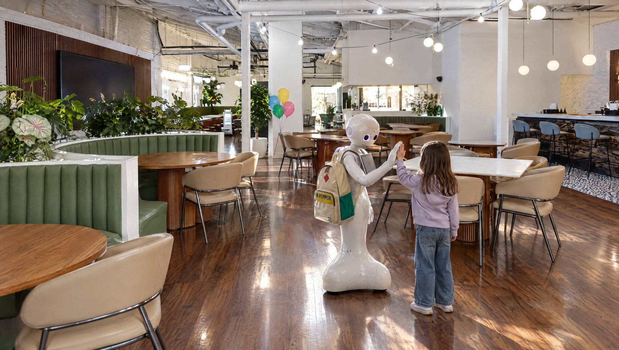 A girl high-fiving Pepper the humanoid robot in the Wonder Studios room, party balloons tied to a chair behind them