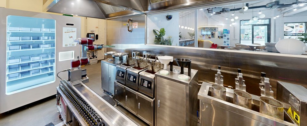 The robot kitchen line seen along the pass: the fridge, the arm on its rail, the fryers and the noodle baths
